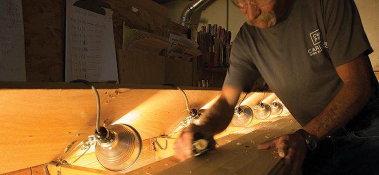 Man sanding wood on a well-lit workbench in a professional woodworking workshop.