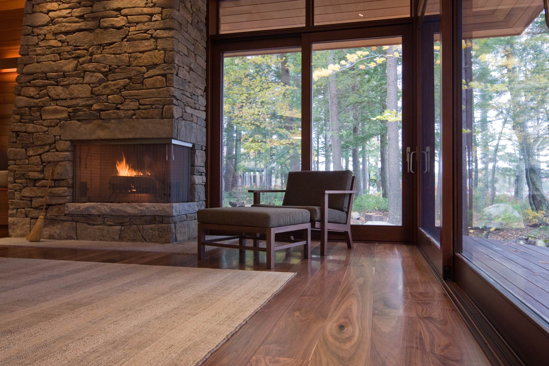 Cozy armchair by a fireplace on wide plank flooring with forest views.